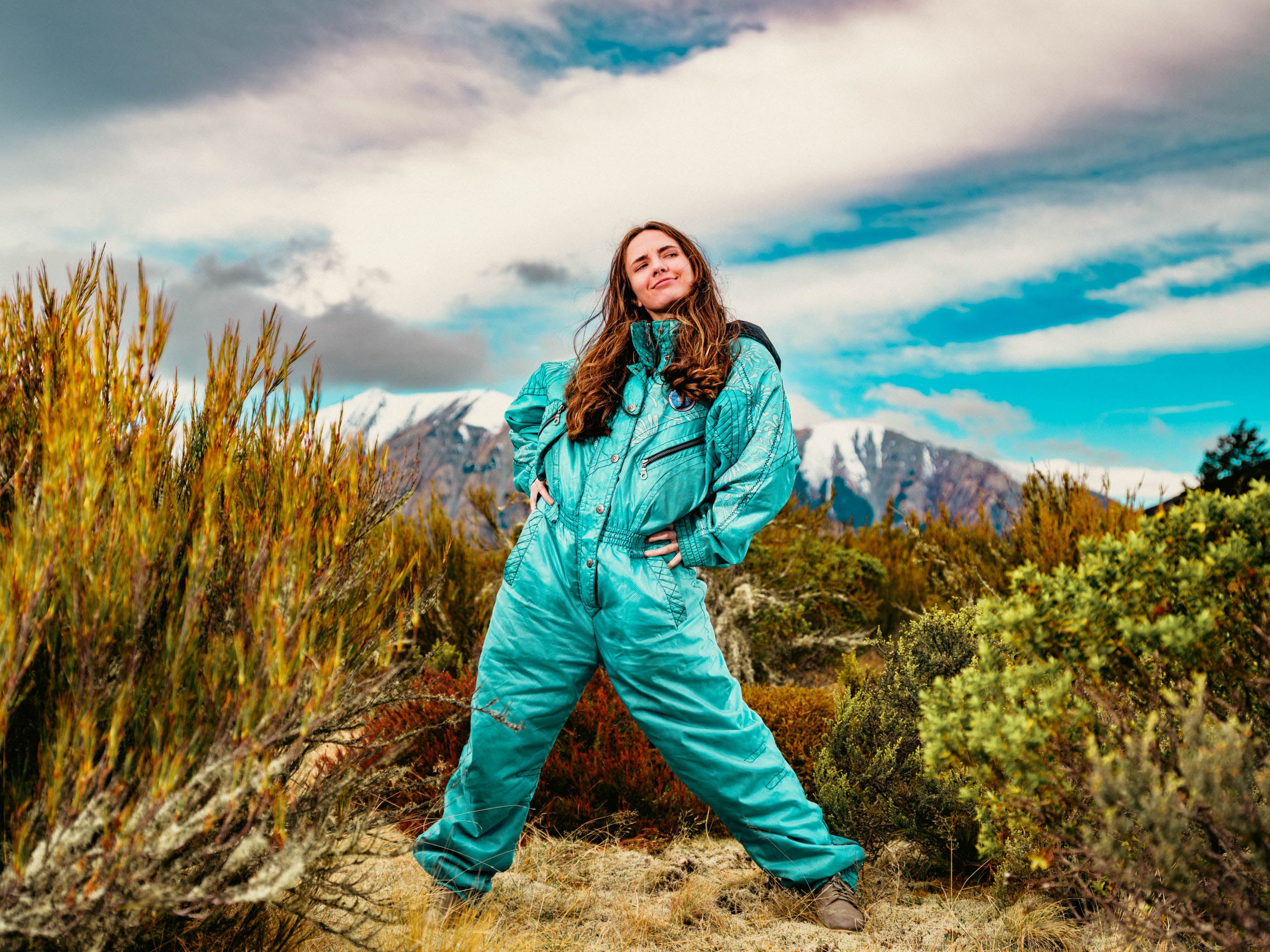 Woman in a turquoise snowsuit standing in a mountainous landscape with clouds and mountains in the background.